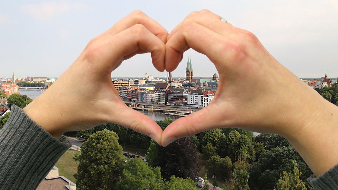 Die Bremer Innenstadt aus der Luft gesehen, davor bilden zwei Hände ein Herz, durch das man die Domtürme sieht.