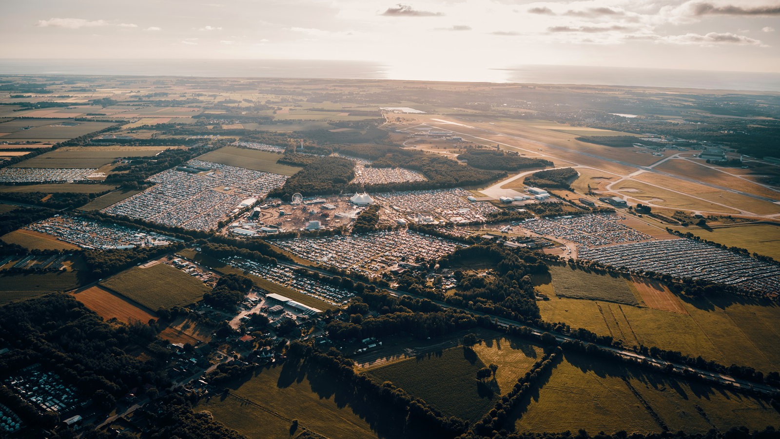 Das aus der Vogelperspektive geschossene Bild zeigt das Deichbrand-Festival in seiner ganzen Größe. Um das Festivalgelände herum sind vollgeparkte Parkanlagen, grüne Wiesen und Baumreihen zu erkennen.