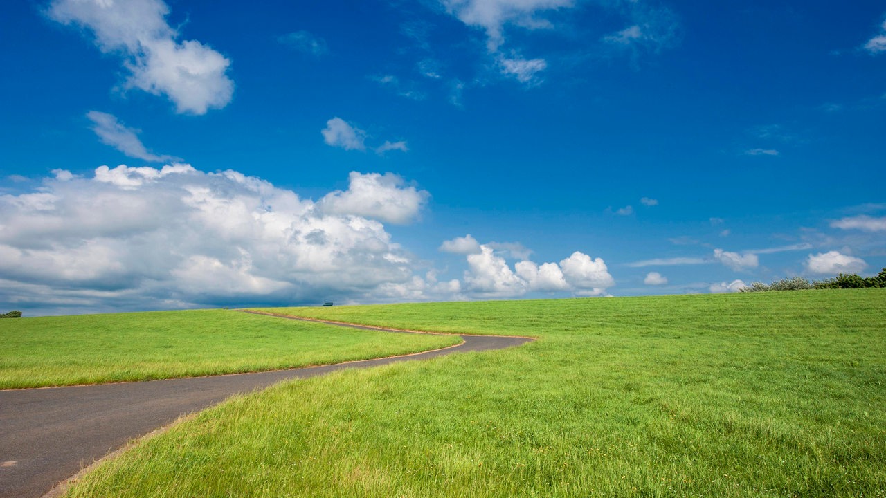 Eine grüne Wiese in der eine geteerte Straße zu sehen ist. Darüber blauer Himmel mit leichten Wolken