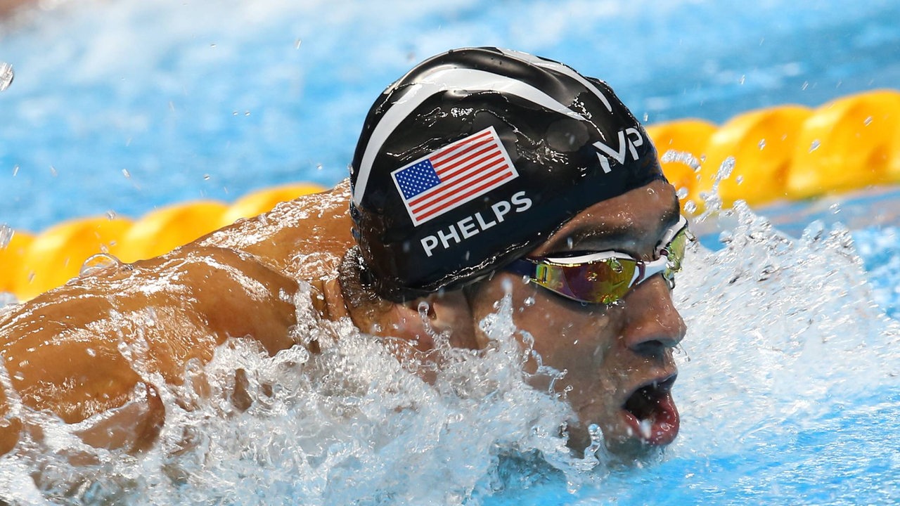 Schwimmer Michael Phelps im Wasser. Er trägt eine Badekappe mit einer USA-Flagge und seinem Namen.