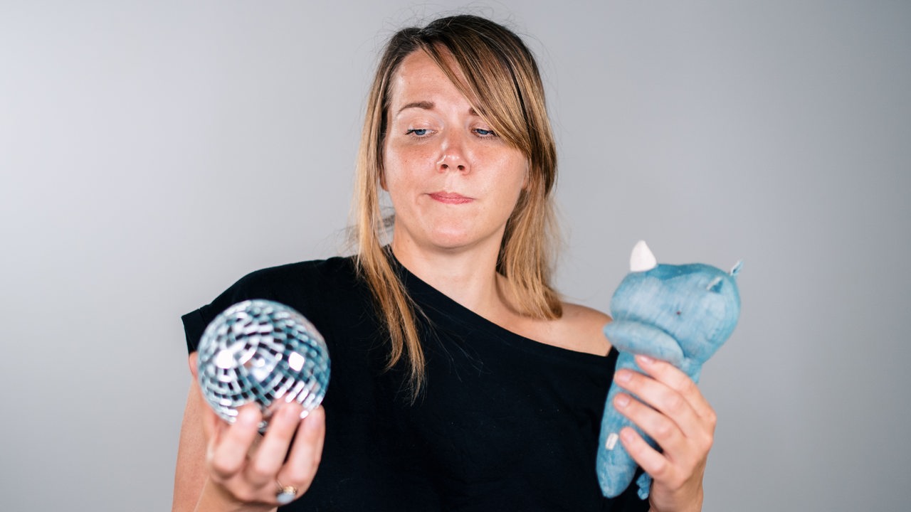 Eine Frau hält in der einen Hand eine Diskokugel und in der anderen ein blaues Kuscheltier, sie schaut skeptisch