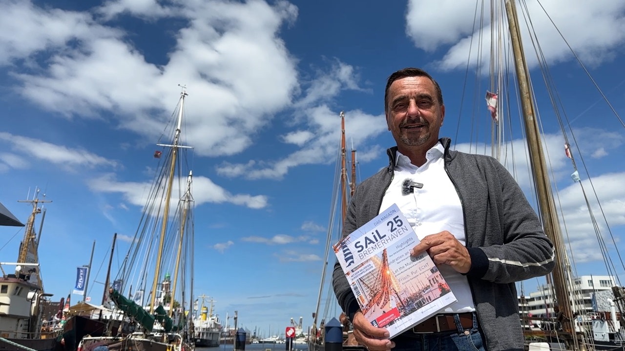 Ein Mann steht vor Schiffsmasten und hält ein Magazin in den Händen.