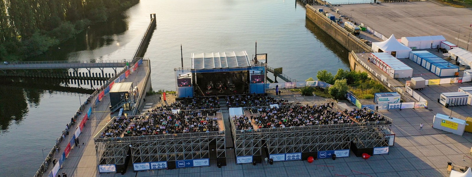 Luftaufnahme einer Open-Air-Bühne mit vollbesetztem Auditorium an einem Hafenbecken