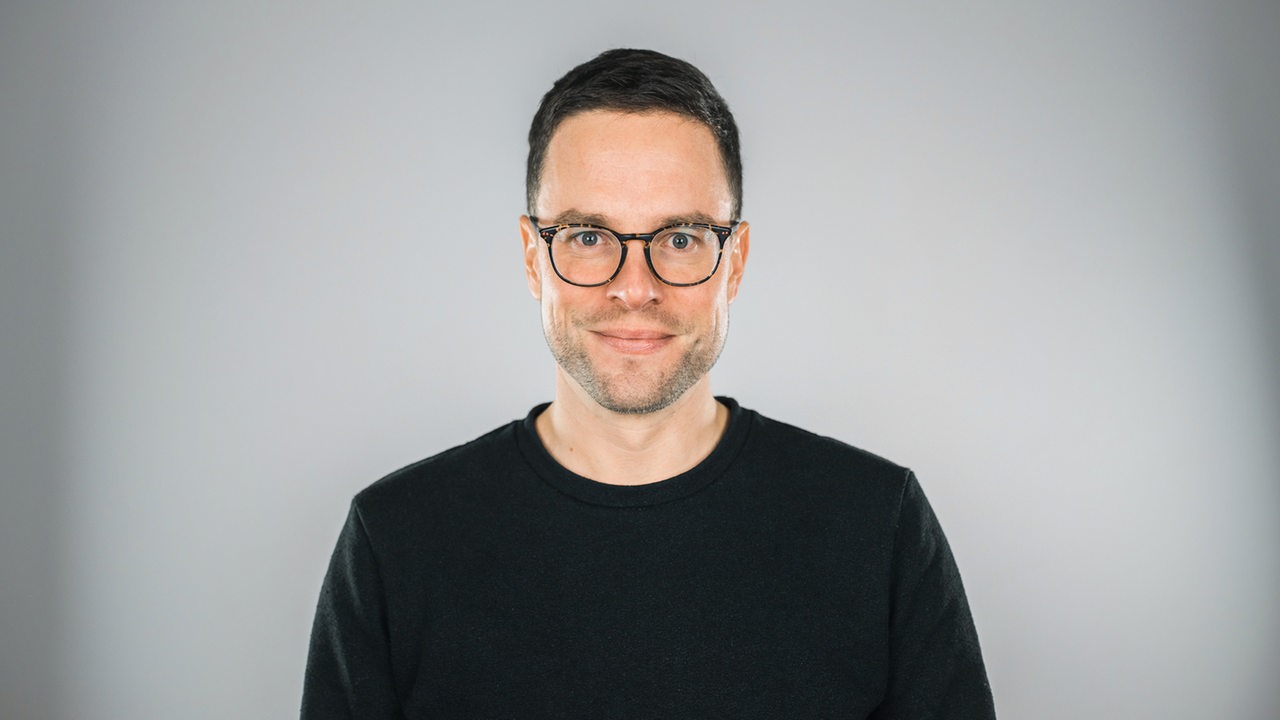 Malte Döbert: ein Mann mit kurzen Haaren und Brille im schwarzen Pullover steht vor grauem Hintergrund
